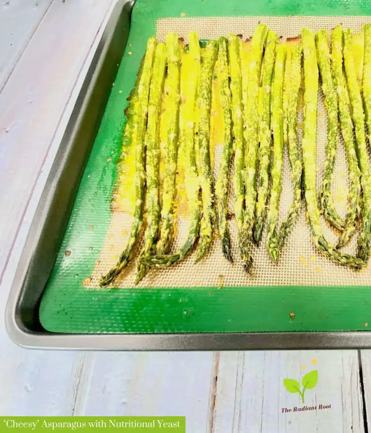 A white wooden table with a baking sheet on top of it. Inside the baking sheet is a green mat and on top of that is roasted asparagus with nutritional yeast “cheese” sauce consisting of nutritional yeast, olive oil, and water. It reads “ ‘Cheesy’ Asparagus with Nutritional Yeast.” | baked asparagus with “cheese”| The Radiant Root