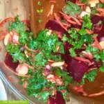 Close up of a clear glass bowl on a wooden table with a tossed and dressed salad containing lupini beans, roasted beets, shredded carrots, and avocado topped with a salad dressing consisting of olive oil, parsley, shallots, lemon zest, dry mustard, and lemon juice. It reads “The Radiant Root” in the bottom left corner. | Lupini beans salad | The Radiant Root