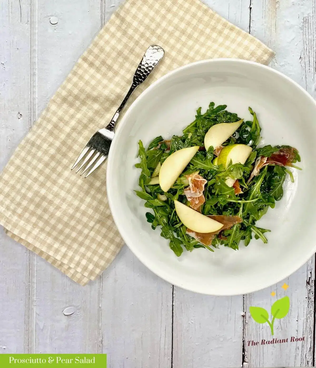 A white wooden table with a brown and white checkered dish towel with a fork on top next to a large white salad bowl containing a pear, arugula, and prosciutto salad with the words "Prosciutto & Pear Salad" in white font with a green highlighting behind it. | Proscuitto Salad | The Radiant Root