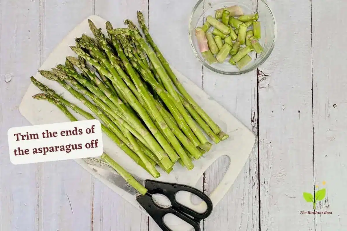 Asparagus with nutritional yeast instruction 3 of 10: A white wooden table with a blue and black marbled cutting board with a pound of asparagus ont top. The ends are cut off and placed inside a small glass bowl. A pair of kitchen shears lay next to the asparagus. It reads “trim the ends of the asparagus off.” | cheesy asparagus recipe | The Radiant Root