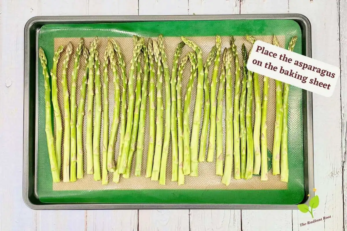 Asparagus with nutritional yeast instruction 6 of 10: A white wooden table with the lined baking sheet containing the asparagus spears arranged in a flat layer. It reads “place the asparagus on the baking sheet.” | cheesy baked asparagus | The Radiant Root