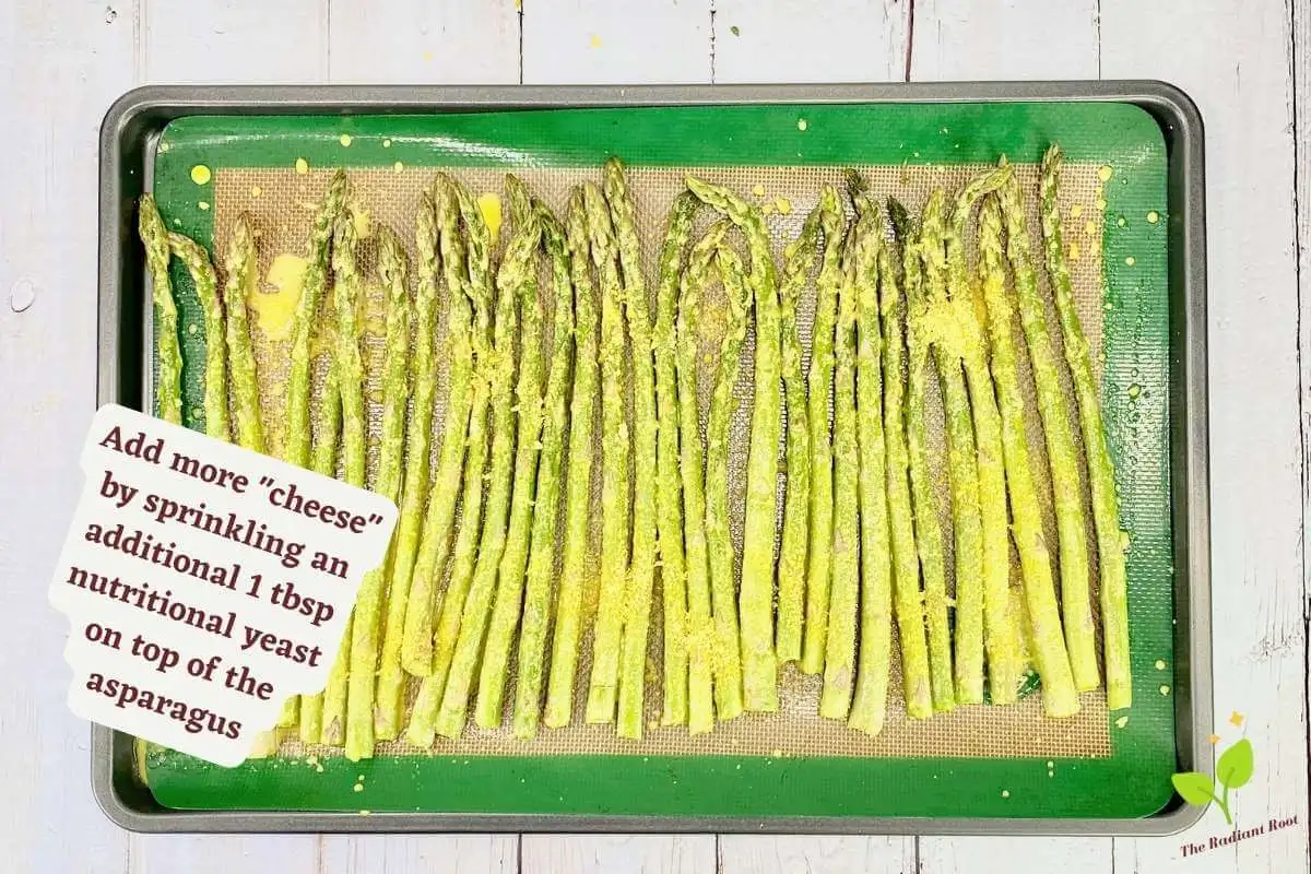 Asparagus with nutritional yeast instruction 8 of 10: A white wooden table with the lined baking sheet containing the asparagus spears covered with “cheese” sauce and the additional nutritional yeast. To the left it reads “Add more "cheese" by sprinkling an additional 1 tbsp nutritional yeast on top of the asparagus.” | asparagus with cheese | The Radiant Root