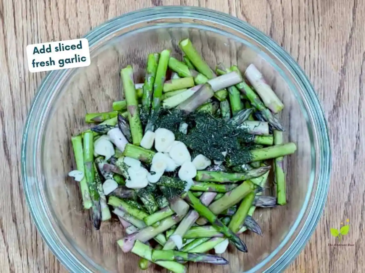 Air Fried Asparagus recipe photo step 4b of 7: A wooden table with a large glass mixing bowl on top of the table containing bite-size asparagus pieces, dried dill, and sliced fresh garlic. It reads “add sliced fresh garlic.” | is air frying healthy | The Radiant Root