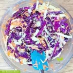 A large clear glass bowl on top of a wooden table. It contains a spatula inside mixed coleslaw with shredded purple cabbage, shredded green cabbage, shredded carrots, coconut cream, apple cider vinegar, and Italian seasoning. It reads “The Radiant Root.” | red cabbage coleslaw | The Radiant Root