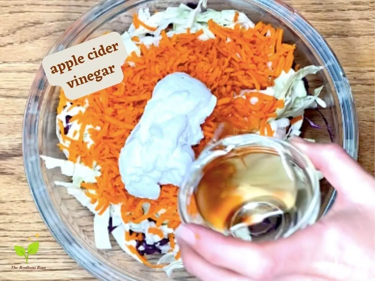 Whole30 coleslaw recipe photo 5 of 8: A wooden table with a large clear glass bowl containing shredded purple cabbage, shredded green cabbage, shredded carrots, coconut cream, and a hand holding a small clear glass dish of apple cider vinegar being poured into the bowl. It reads “apple cider vinegar.” | how to make coleslaw from scratch | The Radiant Root