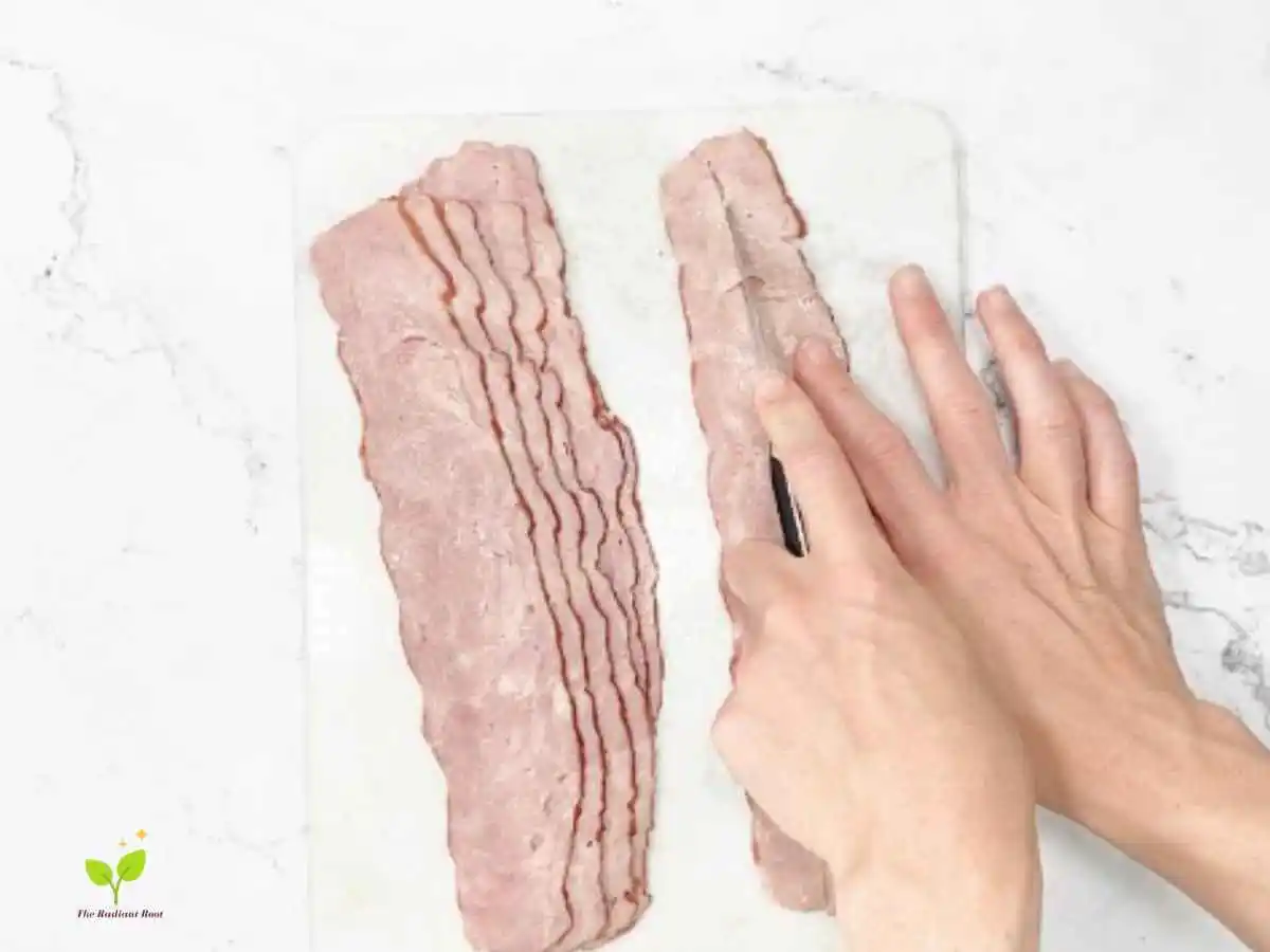 Instructions Photo 3 of 6 for Asparagus Wrapped In turkey Bacon: A white cutting board with hands cutting a piece of turkey bacon lengthwise into two pieces | How long does asparagus take to cook | The Radiant Root