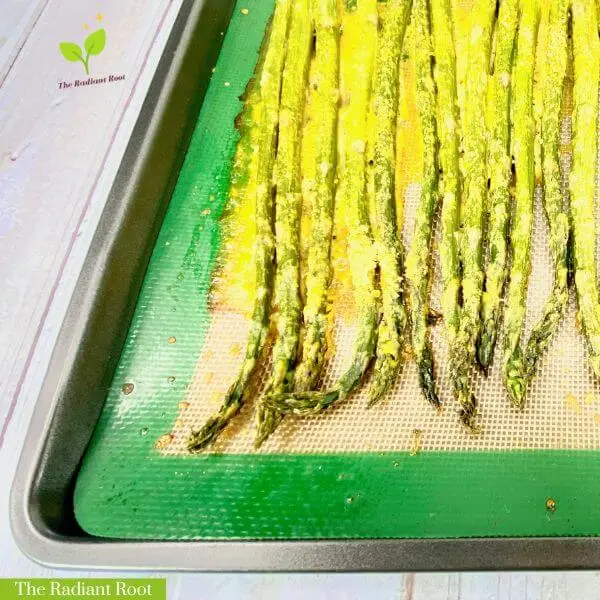 A white wooden table with a baking sheet on top of it. Inside the baking sheet is a green mat and on top of that is roasted asparagus with nutritional yeast “cheese” sauce consisting of nutritional yeast, olive oil, and water. It reads “ ‘Cheesy’ Asparagus with Nutritional Yeast.” | baked asparagus with “cheese”| The Radiant Root