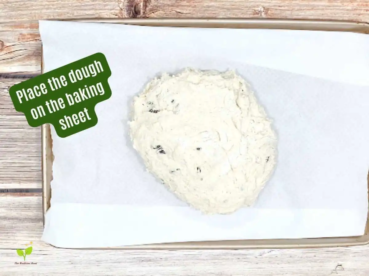 Gluten Free Irish Soda Bread Instruction 10 of 14: The raw soda bread dough loaf on a parchment paper lined baking sheet on the brown wooden table with the words “Place the dought onto the baking sheet.” | Gluten Free Irish Soda Bread | The Radiant Root