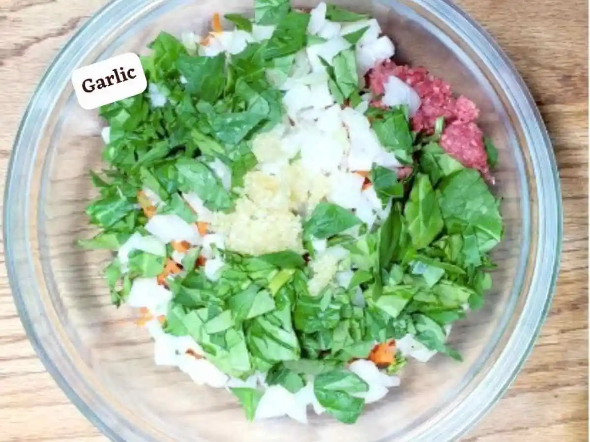 Spinach Meatball photo 6 of 13: A wooden table with a medium clear glass mixing bowl containing grass-fed ground beef, carrots, diced white onion, spinach, and garlic. It reads “garlic” | beef meatball recipe | The Radiant Root