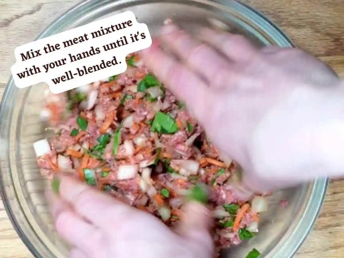 Spinach Meatball photo 9 of 13: A wooden table with a medium clear mixing glass bowl containing the spinach meatball ingredients grass-fed ground beef, carrots, diced white onion, chopped fresh spinach, basil, and oregano being mixed by hands. It reads “Mix the meat with your hands until it’s well blended.” How to make homemade meatballs | The Radiant Root