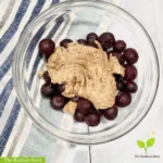 Tigernut butter main photo: A close up of the finished homemade tiger nut butter on top of blueberries in a clear glass bowl sitting on top of a blue striped dish towel. In the lower left hand corner it reads “the radiant root.” | substitute for nuts | The Radiant Root