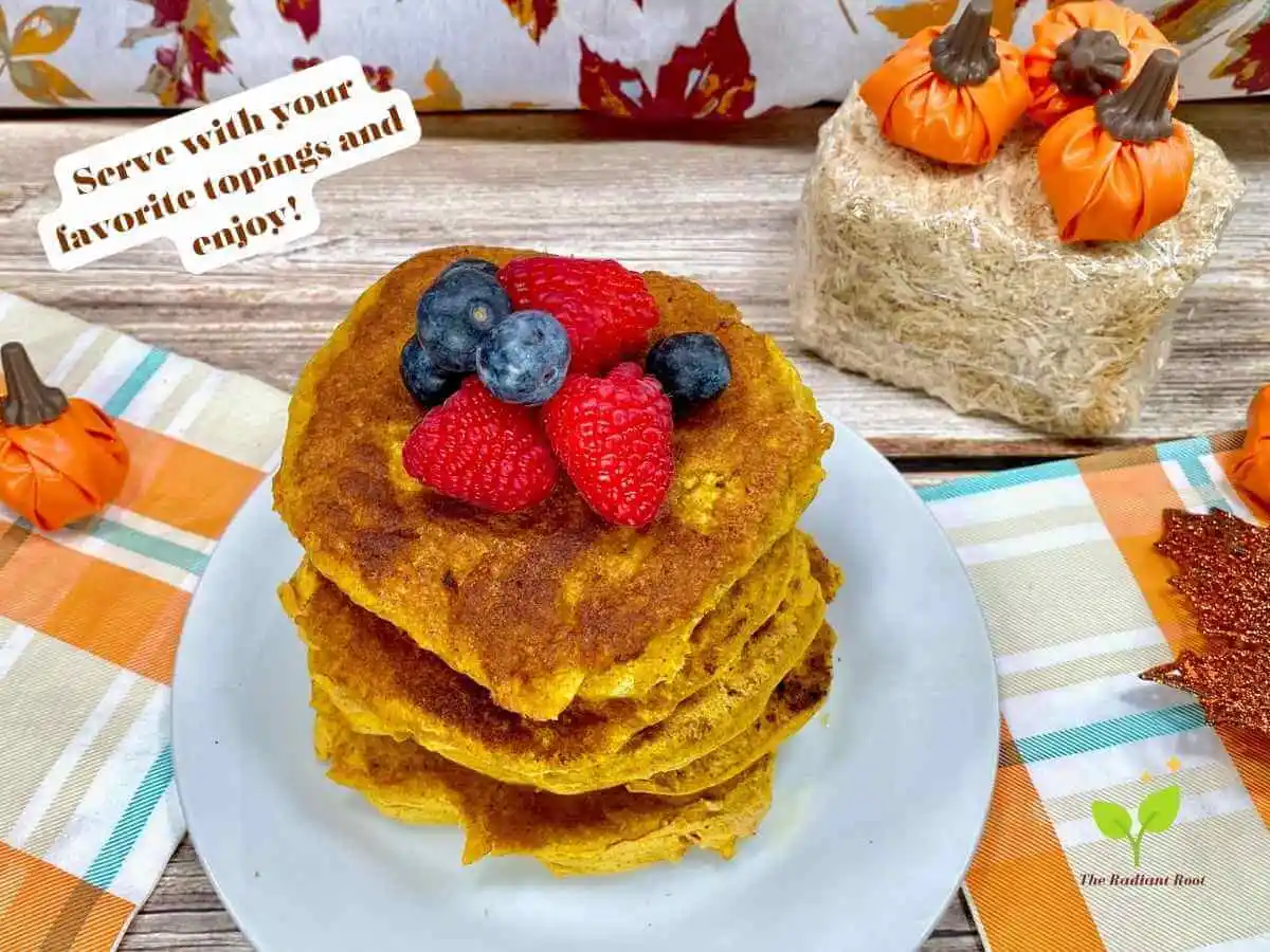 Gluten Free Pumpkin Pancakes recipe instruction photos 12 of 12: A close-up photo of the finished gluten free pumpkin pancakes in a stack on a white plate topped with blueberries and raspberries with a fall backdrop showing fall leaves. Above the pancakes to the left it reads “serve with your favorite topping and enjoy.” | pumpkin pancakes recipe (gluten free) | The Radiant Root