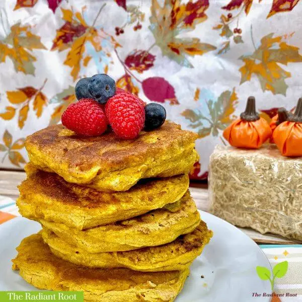 At the top it reads “The Radiant Root.” A close-up photo of the finished gluten free pumpkin pancakes in a stack on a white plate topped with blueberries and raspberries with a fall backdrop showing fall leaves. Below is says “Gluten-Free Pumpkin Pancakes.” | gluten free pancakes | The Radiant Root