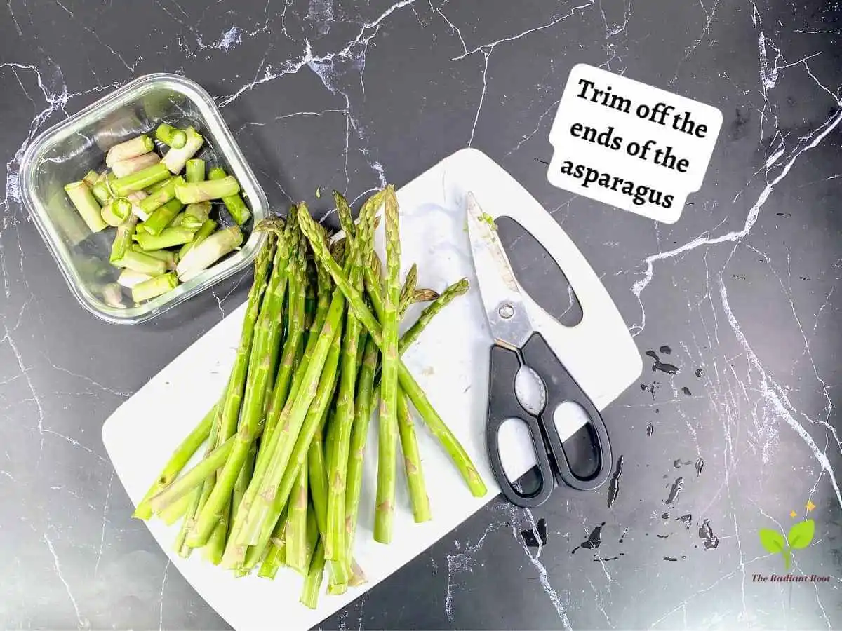 Asparagus with Shallots 1 of 12 : A black marble counter top with a white cutting board containing trimmed asparagus on top of the cutting board. Next to the asparagus is a pair of kitchen shears. In the upper left corner there is a clear glass bowl of the woody ends of the asparagus. It reads “Trim off the ends of the asparagus.” | types of shallots | The Radiant Root