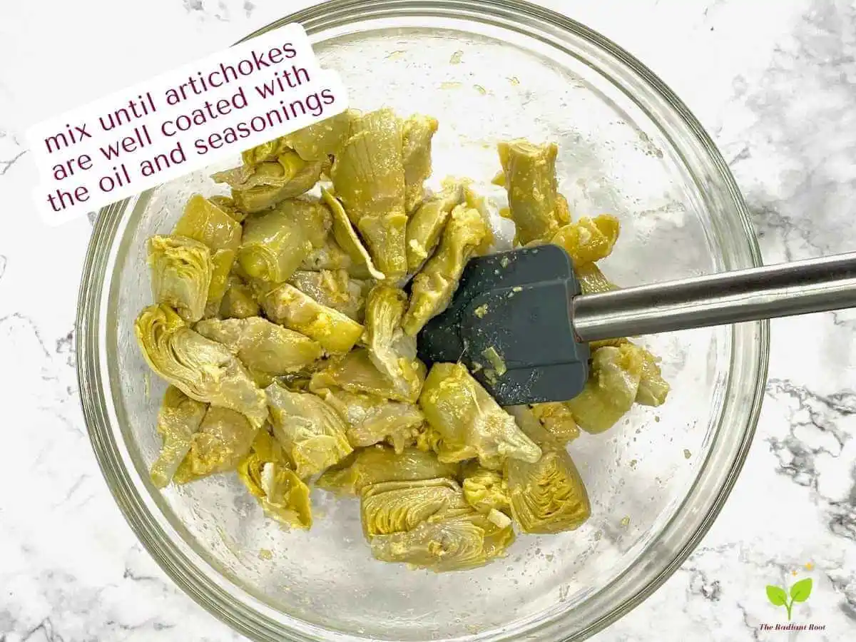 Frozen Artichoke Hearts Recipe instruction photo 8 of 12 : A white marble table with a large clear glass bowl containing thawed artichokes, olive oil, garlic powder, and fresh garlic mixed together with a spatula sitting in the bowl. It reads “mix until artichokes are well coasted with the oil and seasonings.” | appetizers with artichokes | The Radiant Root