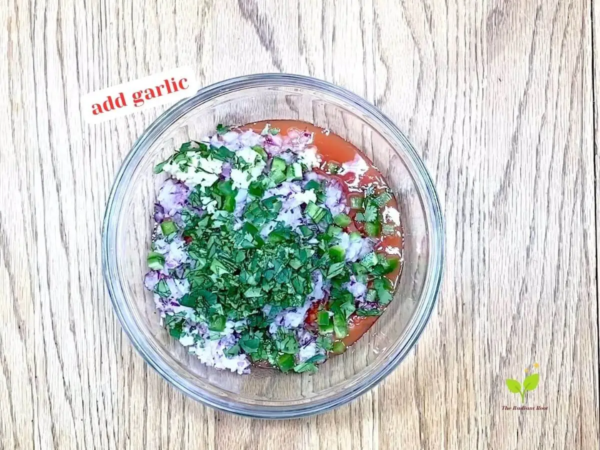 Cilantro salsa recipe instruction photo 5 of 8: A close-up of a wooden table containing a medium glass mixing bowl containing diced canned tomatoes, diced red onion, diced jalapeno peppers, cilantro, and garlic. It reads “Add garlic.” | salsa ingredient list | The Radiant Root