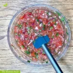 Cilantro Salsa WP Recipe Photo: A close-up of a wooden table containing a medium clear glass mixing bowl with salsa inside the bowl is a blue spatula. In the bottom left corner, it reads “The Radiant Root.” | salsa cilantro lime | The Radiant Root