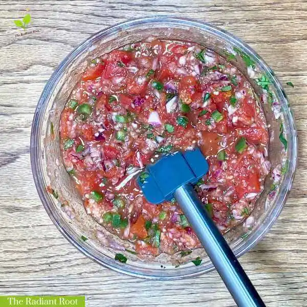Cilantro Salsa WP Recipe Photo: A close-up of a wooden table containing a medium clear glass mixing bowl with salsa inside the bowl is a blue spatula. In the bottom left corner, it reads “The Radiant Root.” | salsa cilantro lime | The Radiant Root
