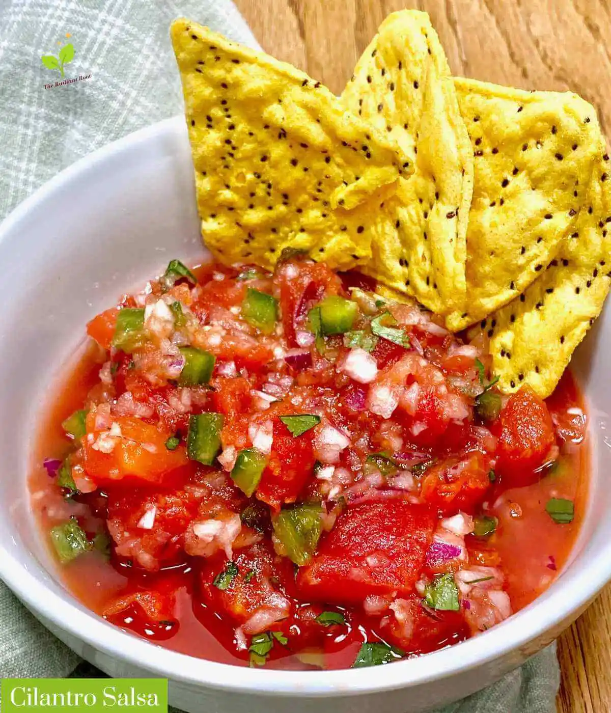 Cilantro salsa main photo: A close-up of a wooden table with a white bowl containing the finished cilantro lime salsa with canned diced tomatoes, red onion, garlic, cilantro, and jalapeno peppers. In the lower-left corner, it reads “Cilantro Salsa” | gluten free salsa | The Radiant Root