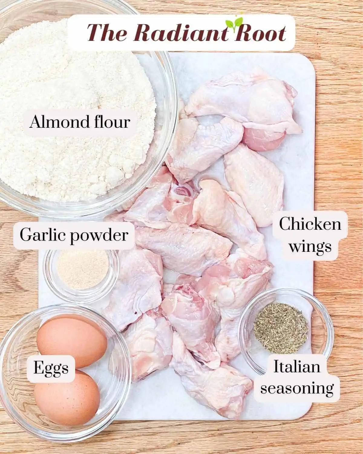 GF Chicken Wings Ingredient photo: A wooden table with a marble cutting board containing raw chicken wings. On the upper left there is a bowl of almond flour. Below the almond flour is a small glass dish of garlic powder. In the lower left corner is a small clear glass bowl of 2 eggs and in the lower right corner it a small clear glass dish of Italian seasoning | alternative to almond flour | The Radiant Root