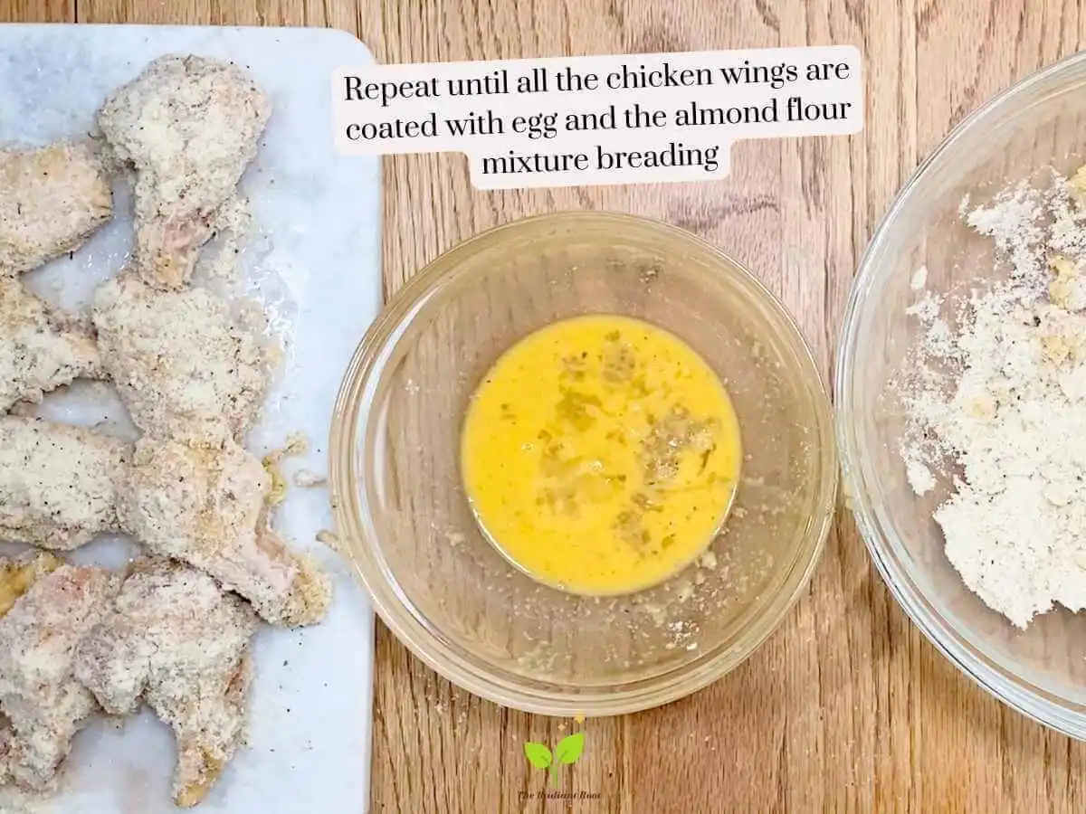 GF Chicken Wings recipe instruction photo 11 of 14 : A wooden table containing three items. On the left is a white marble cutting board containing raw chicken wings coated in the almond flour mixture. In the middle is the medium bowl of scrambled eggs. On the right you see a slight part of the large mixing bowl containing the almond flour, garlic powder, and Italian seasoning mixture. It reads “repeat until all the chicken wings are coated with egg and the almond flour mixture breading.” | reheat chicken wings | The Radiant Root