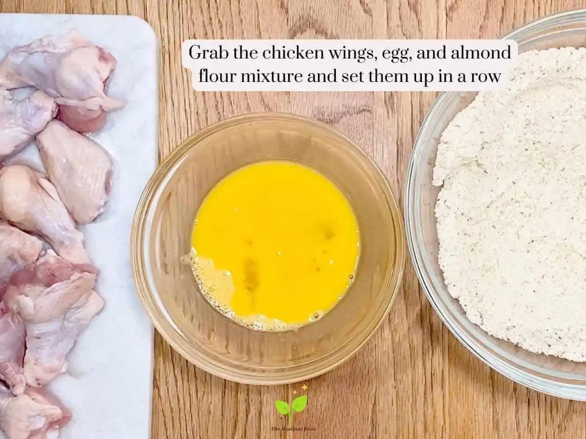 GF Chicken Wings recipe instruction photo 8 of 14 : A wooden table containing three items. On the left is a white marbel cutting board containing raw chicken wings. In the middle is the medium bowl of scrambled eggs and on the right is the large mixing bowl containing the almond flour, garlic powder, and Italian seasoning mixture. It reads “grab the chicken wings, egg, and almond flour mixture and se them up in a row.” | Almond flour nutrition | The Radiant Root