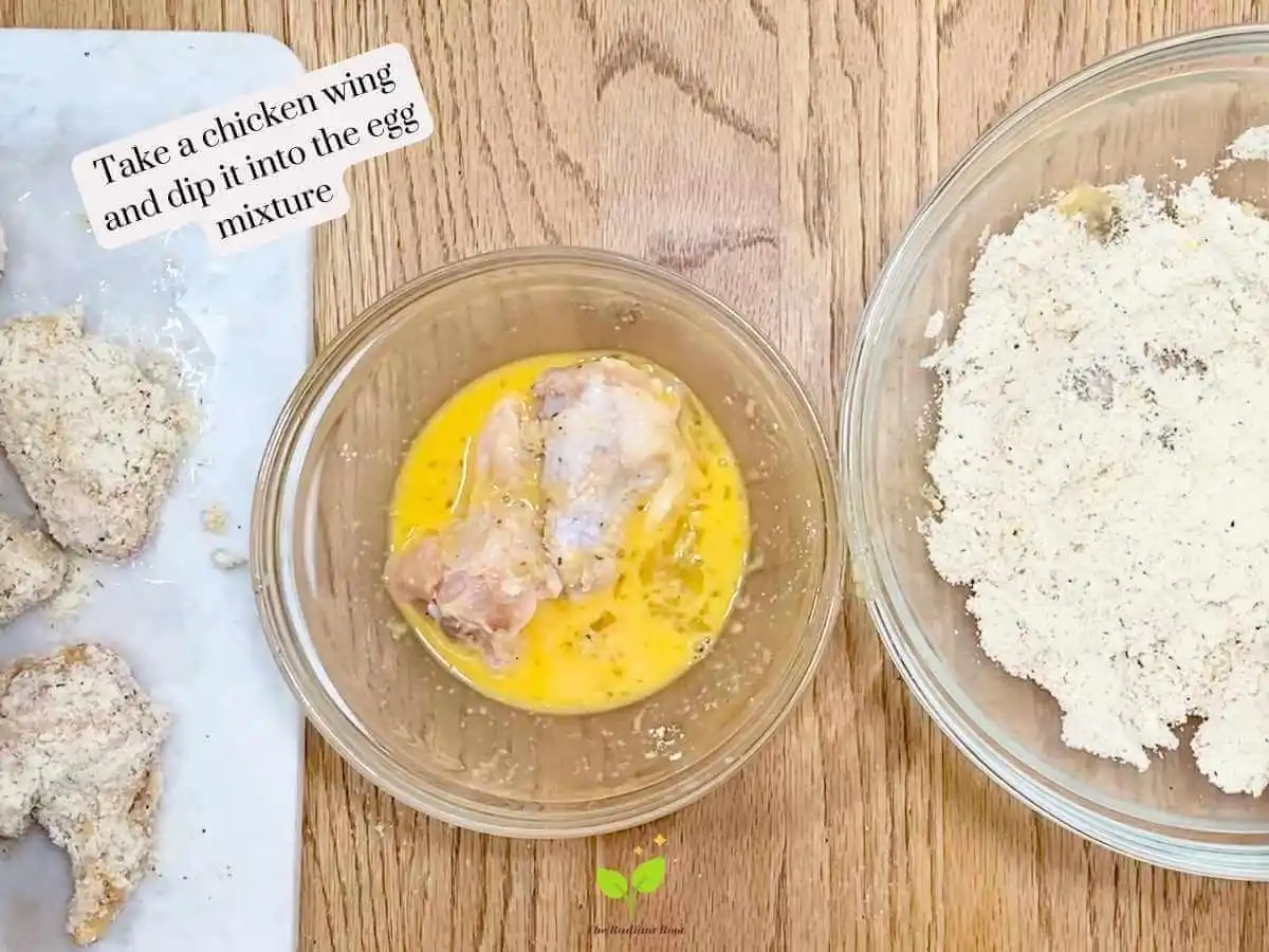 GF Chicken Wings recipe instruction photo 9 of 14 : A wooden table containing three items. On the left is a white marble cutting board containing raw chicken wings. In the middle is the medium bowl of scrambled eggs with two chicken wings sitting in the bowl. On the right is the large mixing bowl containing the almond flour, garlic powder, and Italian seasoning mixture. It reads “take a chicken wing and dip it into the egg mixture.” | baked chicken wings nutrition | The Radiant Root