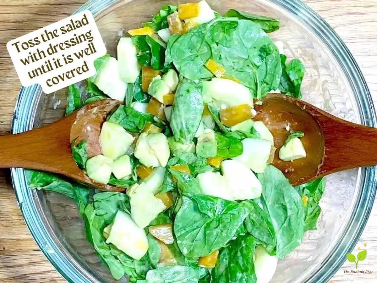 Golden Beet Salad recipe instruction photo 10 of 11 : A wooden table with a close up of a clear medium mixing bowl containing spinach, cucumbers, avocado, cucumber tossed with Dijon dressing, In the bowl are wooden tongs. In the upper left corner it reads “Toss the salad with dressing until it is well covered.” | How to prepare golden beets | The Radiant Root