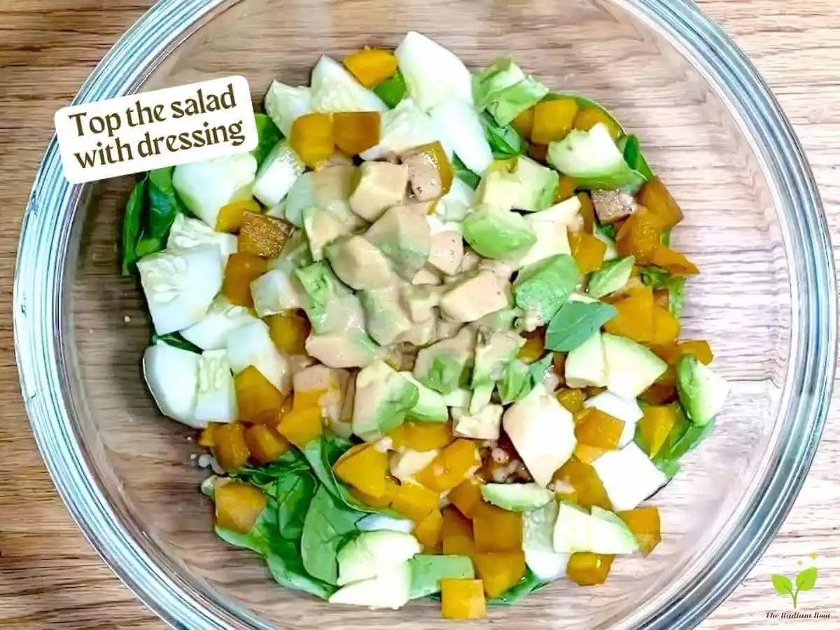 Golden Beet Salad recipe instruction photo 9 of 11 : A wooden table with a close up of a clear medium mixing bowl containing spinach, cucumbers, avocado, cucumber and is topped with Dijon dressing. In the upper left corner it reads “Top the salad with dressing.” | beets health benefits | The Radiant Root