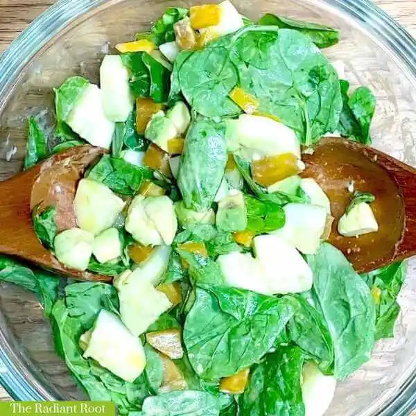 Golden Beet Salad WP Recipe Photo: The close up of a clear glass bowl on a wooden table containing spinach beet salad, cucumber, avocado topped with with a dijon mustard dressing and tongs inside the bowl. In the bottom left corner it reads “The Radiant Root.” | Yellow beets | The Radiant Root