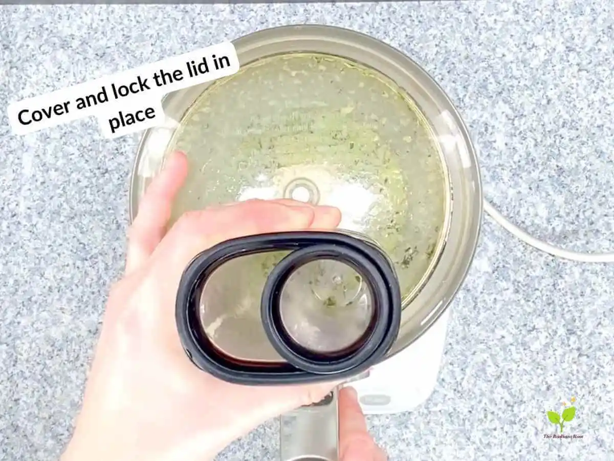 Watermelon avocado salad recipe instruction photo 10 of 20: There is a black speckled table top with a food processor with the food processor bowl on top of it. In the food processor bowl is extra virgin olive oil, cilantro, and lime juice blended and covered with the lid locked in place. In the upper left hand corner it reads “Cover and lock the lid in place.” | how to pick out a watermelon | The Radiant Root