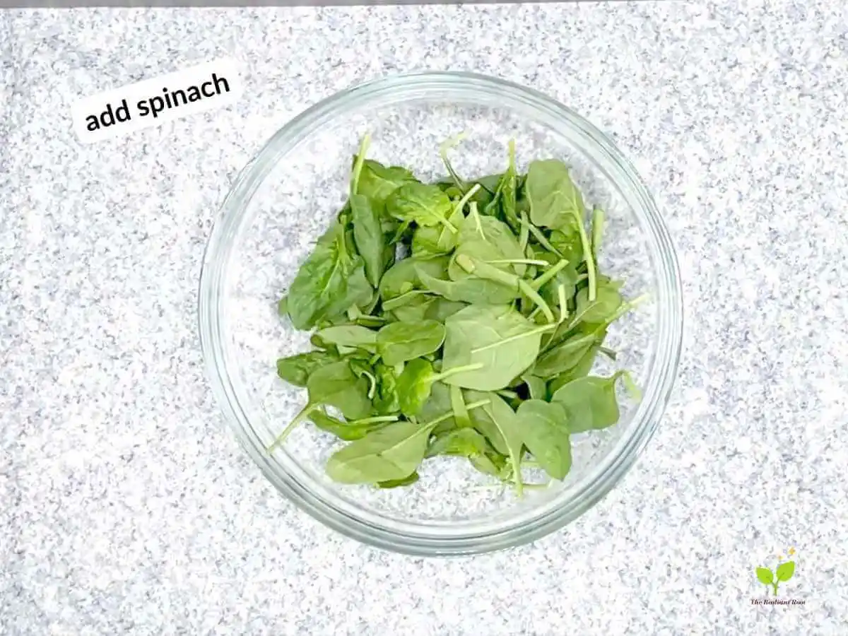 Watermelon avocado salad recipe instruction photo 13 of 20: There is a black speckled table top with a large clear glass mixing bowl on top. Inside the bowl is spinach. In the upper left hand corner it reads “add spinach.”| best time to buy watermelon | The Radiant Root