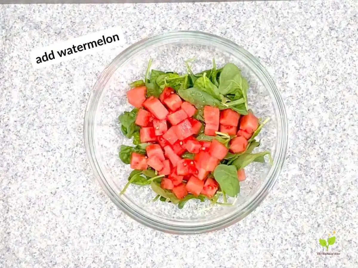 Watermelon avocado salad recipe instruction photo 14 of 20: There is a black speckled table top with a large clear glass mixing bowl on top. Inside the bowl is spinach and watermelon. In the upper left hand corner it reads “add watermelon | how to buy a watermelon | The Radiant Root