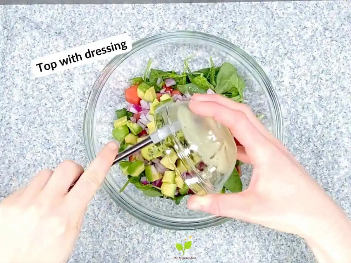 Watermelon avocado salad recipe instruction photo 17 of 20: There is a black speckled table top with a large clear glass mixing bowl on top. Inside the bowl is spinach, watermelon, red onion, and avocado. There are a pair of hands over the bowl pouring the dressing into the salad from a small glass dish using a spatula. In the upper left hand corner it reads “top with dressing.” | what nutrients are in watermelon | The Radiant Root