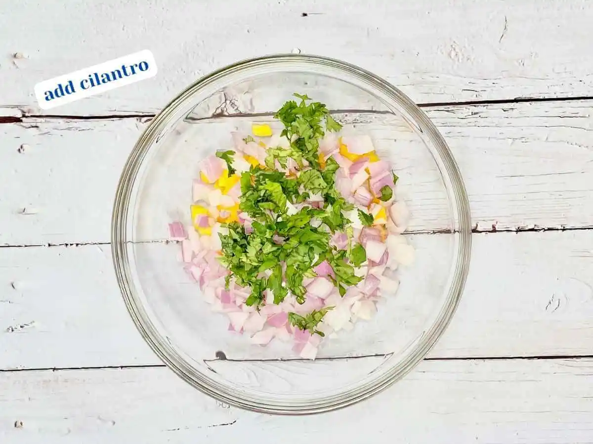 Mango Salsa recipe instruction photo 4 of 7 : A wooden table with a medium clear glass mixing bowl containing diced mango, red onion, and cilantro. In the upper left hand corner it reads “add cilantro.” | How to make mango salsa | The Radiant Root