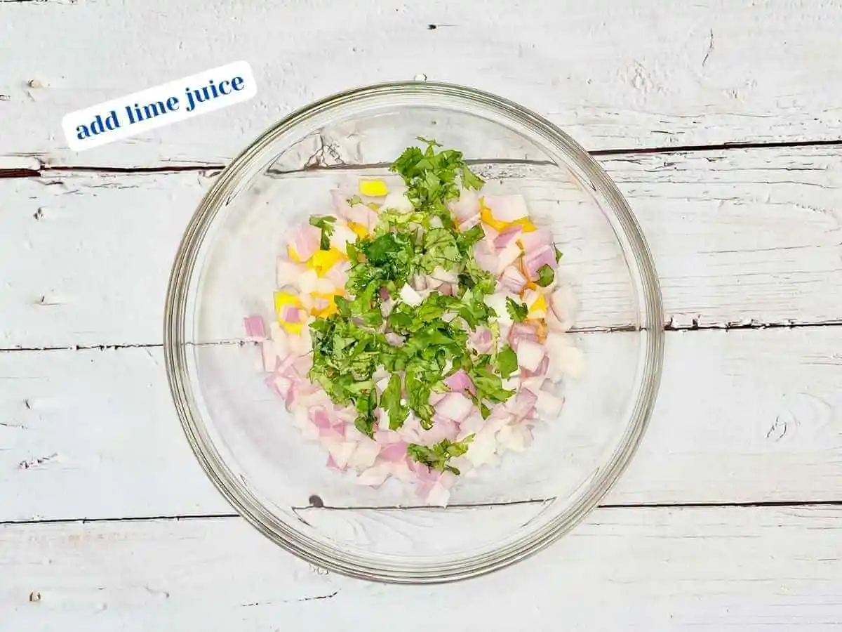 Mango Salsa recipe instruction photo 5 of 7 : A wooden table with a medium clear glass mixing bowl containing diced mango, red onion, and lime juice. In the upper left hand corner it reads “add lime juice.” | What does mango salsa go with | The Radiant Root