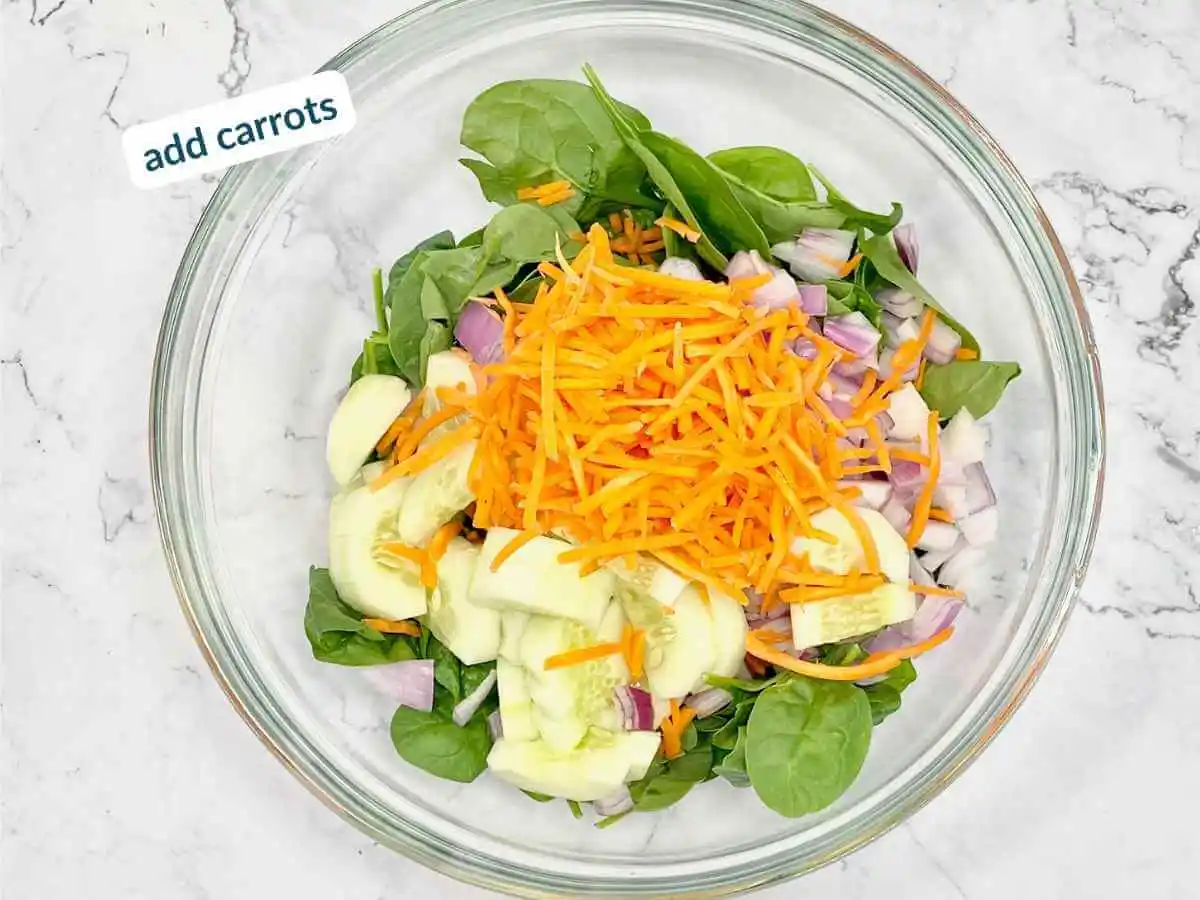 Peach Spinach Salad recipe instruction photo 7 of 12 : A white marble table containing a large clear glass mixing bowl containing spinach, diced red onions, cucumbers, and carrots. In the upper left corner it reads “add carrots.”| salad with peaches | The Radiant Root