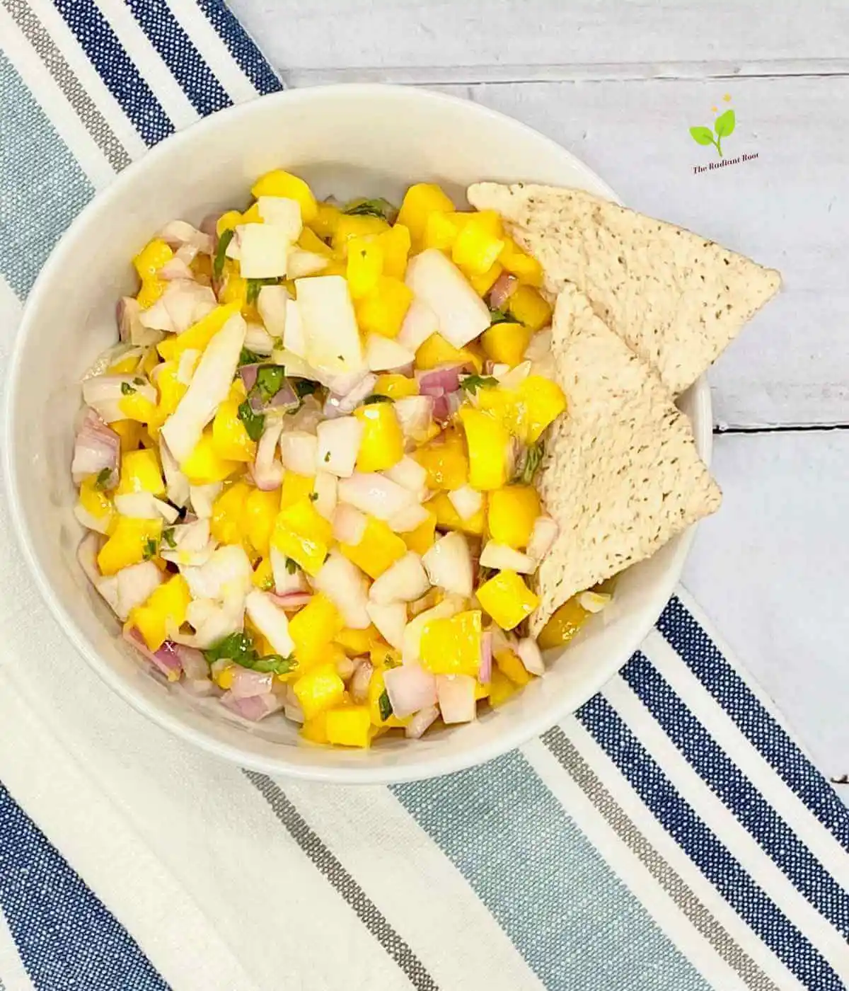 Mango Sala main photo: A close up of a finished mango salsa containing mango, cilantro, red onion, and lime juice in a white bowl on a blue wooden table on top of a blue striped kitchen towel | salsa from scratch | The Radiant Root