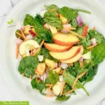 Peach Spinach Salad WP Recipe Photo: A close up of a white marble table containing a large white deep bowl with a finished salad containing peaches, spinach, radishes, cucumber, shredded carrots, and red onion tossed with a red wine vinegar and extra virgin olive oil dressing and topped with sliced peaches as garnish. In the lower left corner it reads “The Radiant Root.” | peach season | The Radiant Root