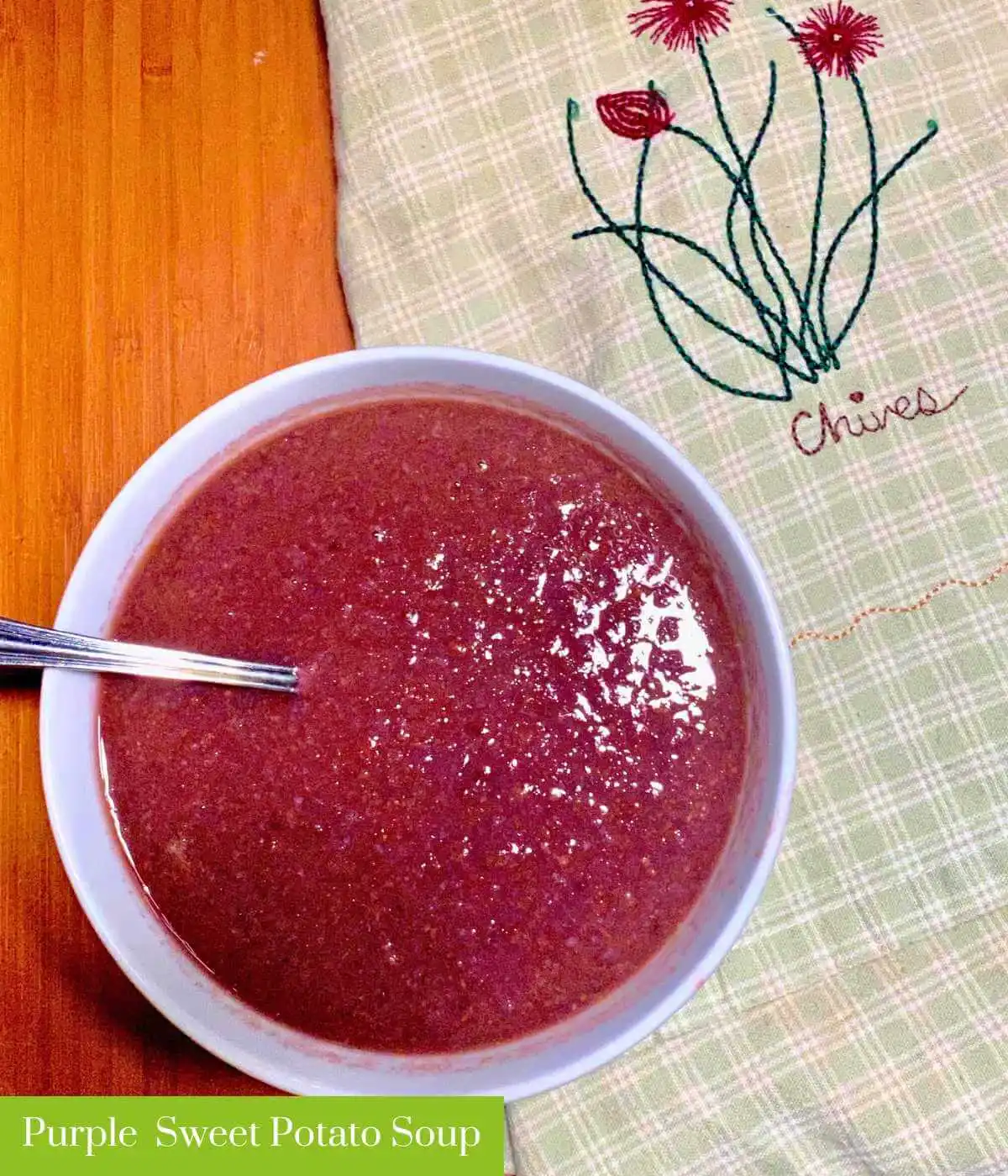 Purple Sweet Potato Soup with Cauliflower in white bowl with spoon next to a dish towel that says "Chives." In the lower left corner it reads "Purple Sweet Potato Soup." | The Radiant Root